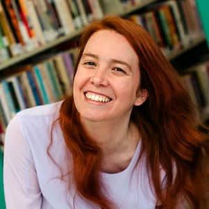 Portrait of Brianna, a woman with light skin and red hair, laughing and leaning sideways with library shelves in the background.